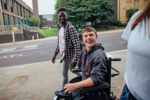 Three students, including one in a wheelchair, laughing together on a sidewalk.