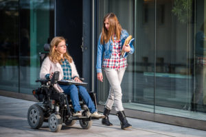 Two university students, one in a wheelchair, on a sidewalk on a college campus.