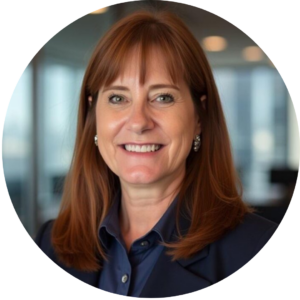 Eve Hill headshot. A professional headshot of a woman with shoulder-length auburn hair, wearing a navy blue blazer and matching blouse. She is smiling and wearing silver earrings. The background features a modern office setting with large windows and blurred lights.